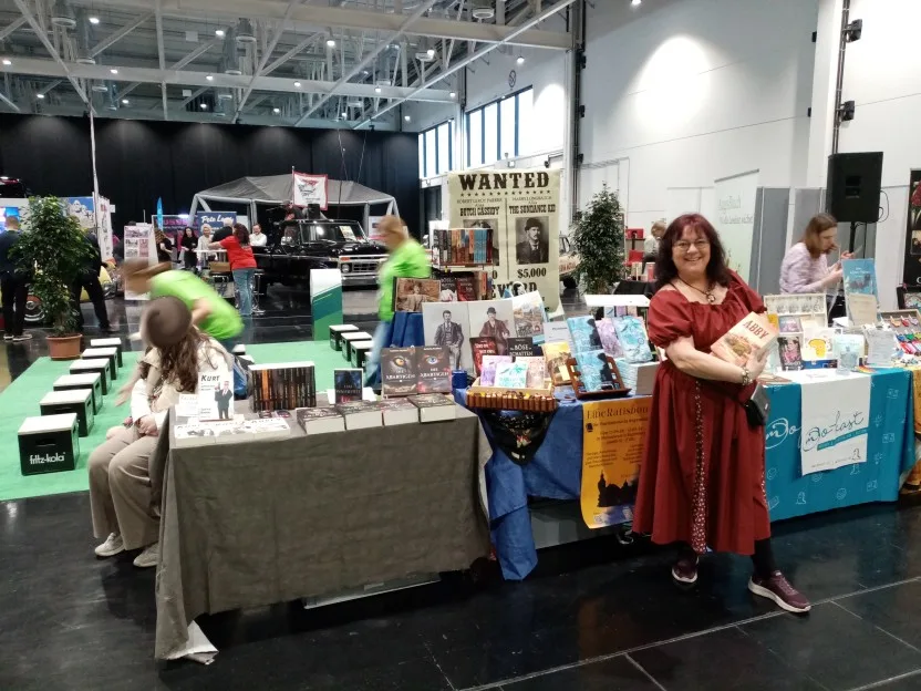 Neben unserem Stand ist der von Claudia, hinter dem Steckbriefe und Photos alter Ganster hängen. Auf dem Tisch ihre Romane, davor sie im roten Kleid, wie sie ihren Roman "Abby" in die Kamera hält. Am Tisch hängt ein Plakat für die LibeRatisbona.