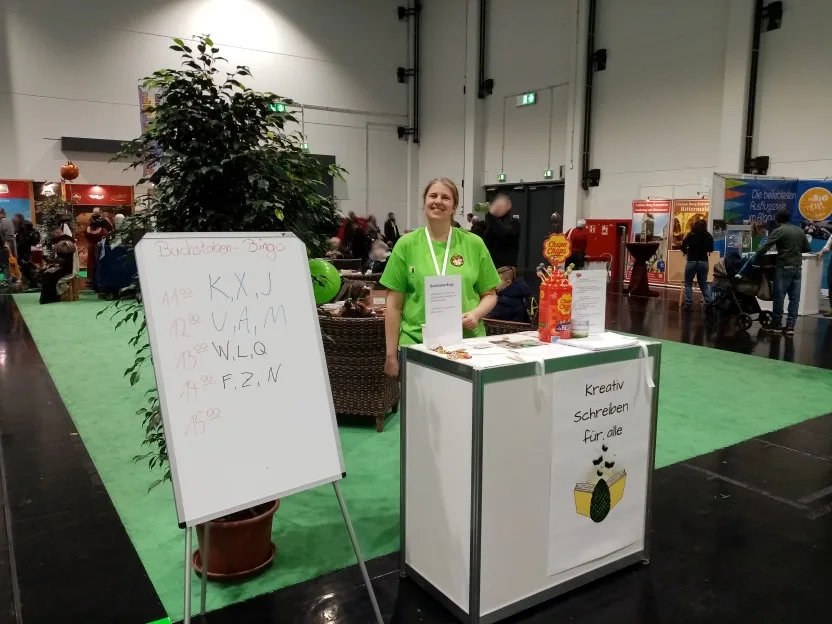 Lucie steht im quietschgrünen T-Shirt hinter einem kleinen Stand, auf dem einige Zettel und etwas Süßkram liegen. Vorne am Stand ein Poster, das für ihre Kurse in kreativem Schreiben wirbt. Sie lacht in die Kamera. Hinter ihr die Sitzgruppen.