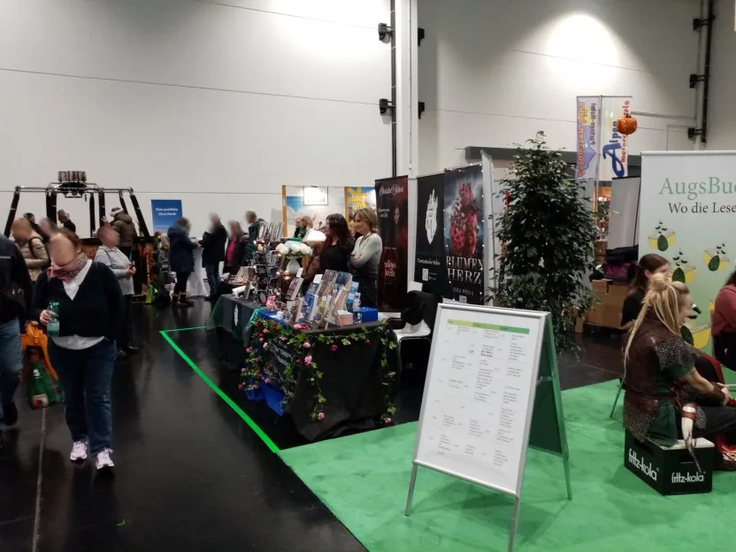 In der Mitte des Bildes ein mit Blumenketten geschmückter Stand, auf dem dunkelblauen Tuch stehen Olkas Bücher. Sie steht hinter ihrem Stand, hinter ihr wiederum ein Banner zu ihrem Buch Blumenherz. Etwas hinter Ilka steht Christel Scher an ihrem Stand, links sind Besucher zu sehen, rechts teilweise die Teilnehmerinnen einer Podiumsdiskussion.