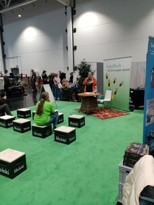 Ivetta steht mit dem Microphon vorne auf dem mit grünem Teppich markierten Lesungs- und Vortrags-Bereich. Für Zuhörer wären Cola-Kästen als Sitzgelegenheiten aufgestellt, aber nur Lucie sitzt da und Ivettas Begleiter am Rand des Bildes.