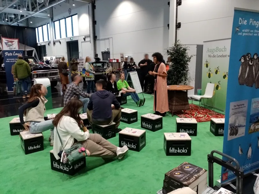 Karima, eine dunkelhäutige, schlanke Frau mit "Afro"-Frisur, steht auf der rechten Seite, vor ihr auf den Zuhörer-Kisten sitzen schon so einige Leute. Noch sind aber die Hälfte der Kisten leer.