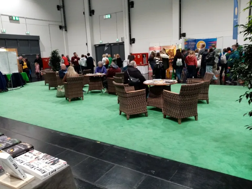 Auf einem grünen Teppich stehen drei Tische mit jeweils drei oder vier Korbsesseln drumrum. Auf den Tischen sieht man die ausgelegten Bücher und Werbematerialien, auf einigen der Sessel sitzen Besucher.