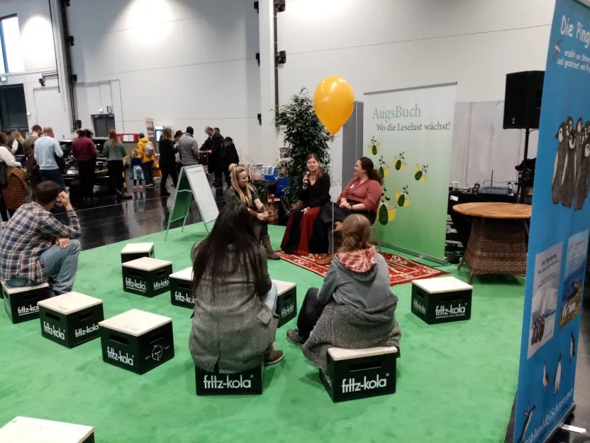 Auf der bekannten grünen Fläche sitzen vorne drei Autorinnen, die linke ist Mira Valentin, die mittlere hält gerade das Mikrophon. Auf den Cola-Kästen sitzen doch immerhin drei Zuhörer.