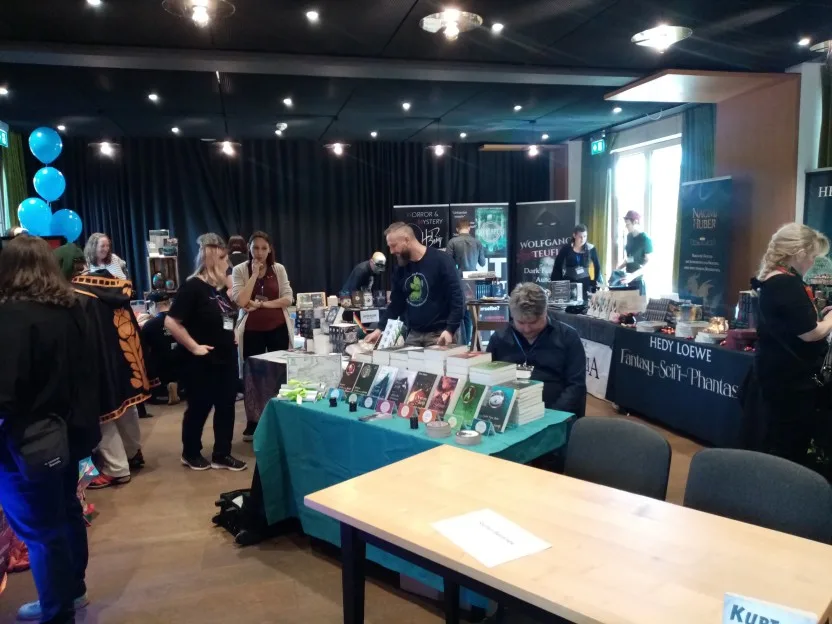 Blick in den Fantasy-Saal.Einige Aussteller sind noch mit dem Aufbau beschäftigt, andere unterhalten sich. Vorne sieht man einen leeren Tisch, daneben ragt mein Kurt ein bisschen ins Bild. Hinten verdeckt ein Vorhang wohl eine Bühne, und jemand hatte für die Kinder blaue Luftballons mitgebracht.