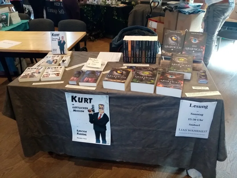 Mein Tisch. Auf der üblichen dunkelgrauen Tischdecke steht links Kurt 1, davor liegen die Teile 2, 3 und 4. Daneben liegt Lia, dann kommen die Sammelbände der Abartigen, von denen ich die Hardcover-Versionen von Band 1 und 3 aufgestellt habe. Die Einzelbände stehen aufgereiht zwischen Buchstützen, und meine Kurzgeschichten liegen auch noch da. Vorne an der Tischdecke hängt ein Plakat von Kurt und eine Ankündigung für meine Lesung am Sonntag Nachmittag. Zwischen den Büchern liegen Lesezeichen und einige ausgedruckte Leserstimmen.