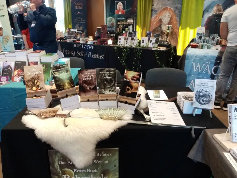 Auf Stefans Tisch mit schwarzer Tischdecke liegen neben einem weißen Lammfell auch ein Schwert und eine Kampfaxt. Seine Bände stehen jeweils auf Stapeln desselben Buches. Daneben liegen Lesezeichen und eine Preisliste. Vor dem Tisch steht ein großes Bild vom Cover seines ersten Reihenbandes.