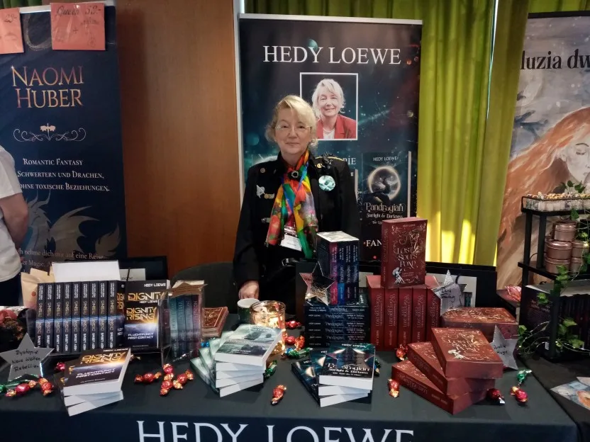 Hedy hinter ihrem Stand. Sie trägt ein schwarze, uniformartige Jacke mit diversen Applikationen, unter anderem einem Sternenflotten-Abzeichen, und ein sehr buntes Halstuch. Vor ihr sind ihre Bücher präsentiert, links SciFi, rechts Fantasy, dazwischen Deko aus bunten Bonbons.