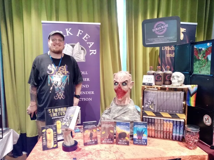 Mark Fear hinter seinem Tisch. Er trägt eine Schirmmütze und ein T-Shirt mit der Aufschrift "Ghost"wobei das T am Ende wie ein umgedrehtes Kreuz aussieht. Sein Tisch wird unter anderem von einer grusligen Maske geziert, so eine Art Zombie mit sehr blutverschmiertem Mund. Dazu eine aufgestellte Hand, die sein neuestes Buch hält, und ein Totenkopf.