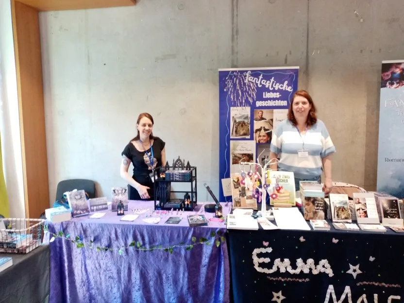 Links Petra Profant, ihr Tisch ist sehr in Lila gehalten. Rechts hat Emma Male eine offenbar selbst gestaltete Tischdecke mit ihrem Namen aus Bordüre-Bändchen und weiteren aufgenähten Verzierungen und einen Aufsteller hinter sich, auf dem "fantastische Liebesgeschichten" steht. Beide stehen hinter ihrem Tisch und lächeln in die Kamera.