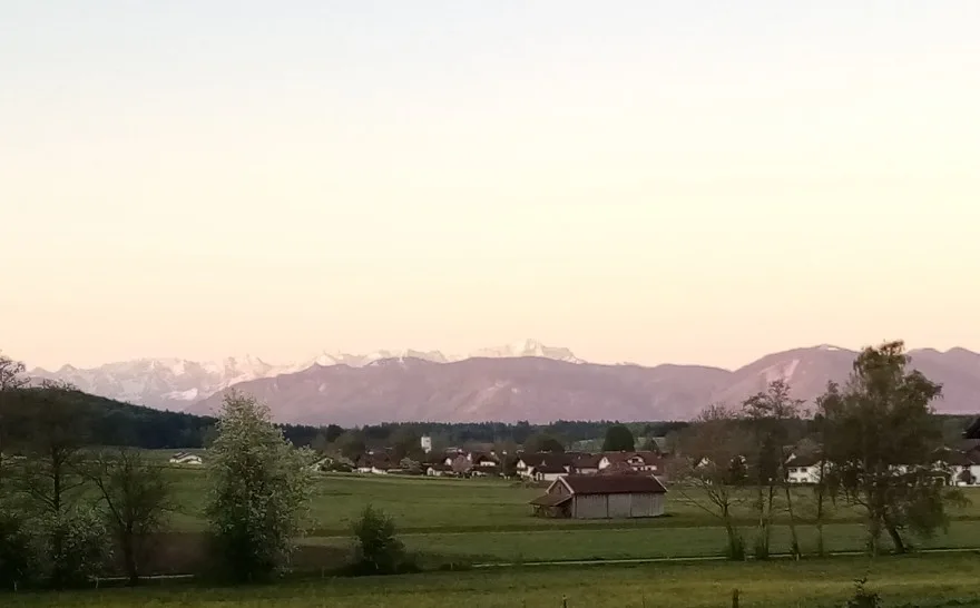 Ein Blick auf die Zugspitze und einen Teil der umgebenden Alpen im Morgenlicht. Der Himmel ist wolkenlos. Im Vordergrund Wiesen, ein paar Bäume und einige Teile des Dorfes, dahinter waldige Hügel.