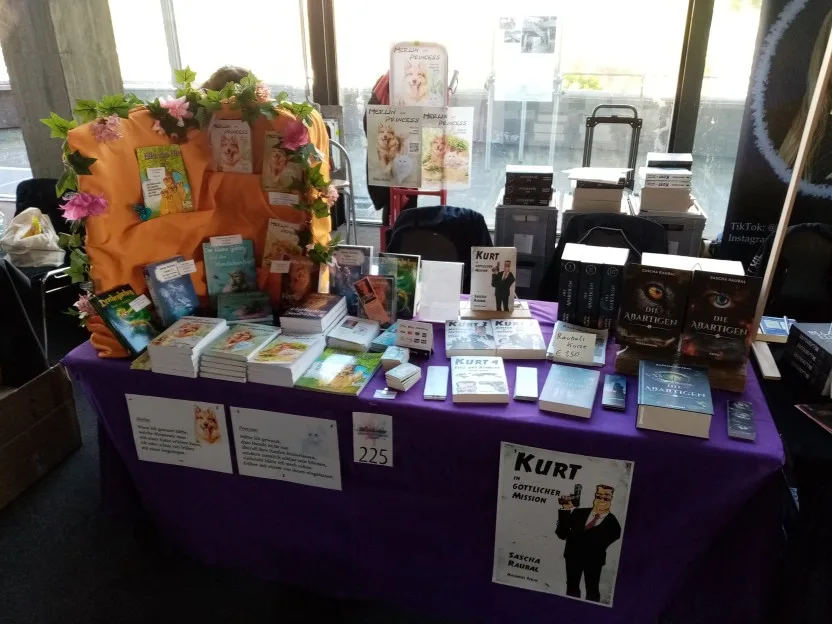 Unser Stand. Auf einer dunkel lila Tischdecke liegen und stehen die Bücher ausgebreitet, dazwischen Lesezeichen usw. Petra hat links noch einen alten Koffer aufgestellt, mit orangenem Tuch überdeckt, und darin Bücher wie in einem Regal präsentiert. Drumherum eine Girlande aus Kunstblumen. Vornedran hängt rechte ein Plakat von meinem Kurt und links kleine Charakterportraits von Hund Merlin ud Katze Princess.