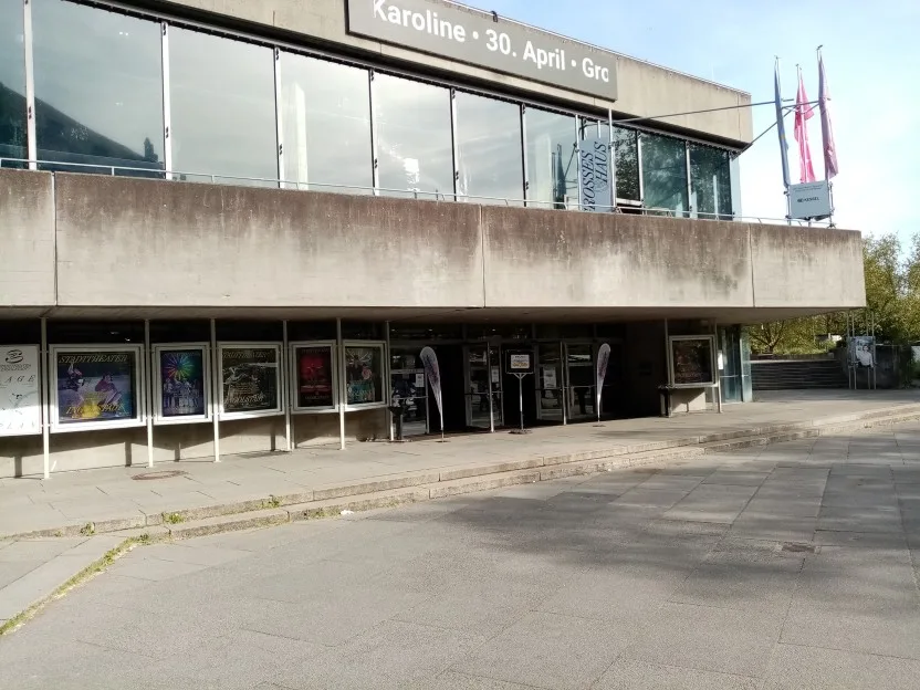 Außenansicht des Stadttheaters. Viel Beton, im ersten Stock eine große Glasfront. Der Beton ist alt und schmutzig. Unten sieht man Glaskästen mit diversen Plakaten und rechts davon den Eingang.